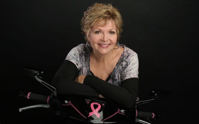 A person is smiling, leaning on a bicycle with a pink ribbon, symbolizing breast cancer awareness, set against a dark background.